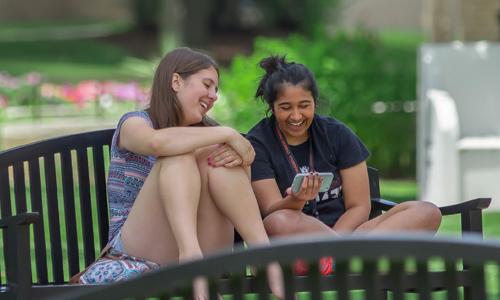 two Carroll students sitting on a bench laughing and looking at a cell phone.